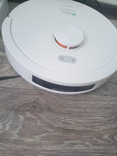 The image shows a white robotic vacuum cleaner resting on a wooden floor. The vacuum is circular with a slightly raised top section that has an orange accent. There are two buttons visible on the top of the vacuum: one with a house icon and another with a power symbol. A dark rectangular sensor is present on the front of the vacuum. The wooden floor has a light brown and gray grain pattern. The scene is indoors, and the lighting is moderate. There is no indication of the city of Podporozhye, Russia in the image.