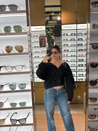 A woman stands in front of a mirror, holding a phone and wearing sunglasses, a dark jacket, and jeans. The mirror reflects a display of sunglasses, with brands like Cartier and Ray-Ban visible. The shelves are filled with eyewear in various styles and colors. The lighting is bright and the overall scene suggests a retail store environment, likely an eyewear or department store.