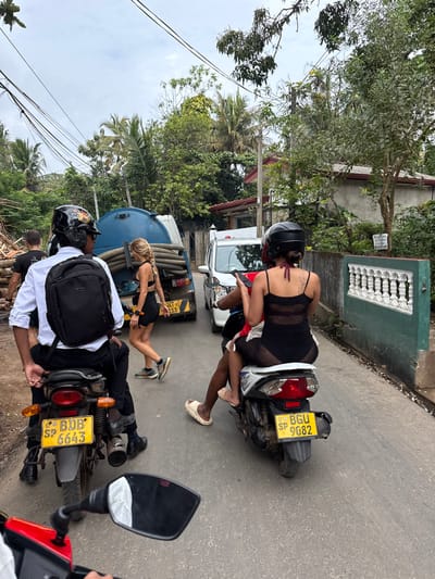 Two motorbikes are parked on the side of a road. Both vehicles have yellow license plates, with one reading "BUB 6643" and the other "BGU SP 9082." A woman in a black mesh dress is sitting on the back of the first motorbike. Nearby, another woman in a black dress stands between the bikes. The drivers are wearing helmets and the first driver has a white shirt. A truck with a blue tank is parked next to them. Trees line the road.