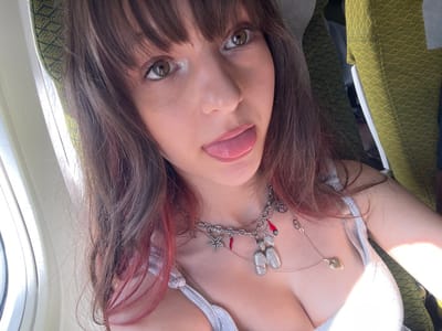 A young adult female is depicted in a close-up selfie taken within an airplane cabin. She possesses long brown hair with reddish/pinkish undertones and bangs. Her eyes appear light brown or hazel. The subject is directly facing the camera with her tongue extended. She is wearing a light-colored, possibly white or light grey, spaghetti strap top that reveals cleavage. Around her neck, she wears multiple layered necklaces. One prominent silver-toned chain features several charms, including a starfish, red chili peppers, and a large, textured pendant resembling two conjoined shapes or the letter 'M'. A second, finer chain with a small heart-shaped pendant is also visible. To the left of the frame, a segment of an airplane window is visible, through which strong daylight enters. The background on the right and top reveals green fabric with a geometric, possibly diamond, pattern, consistent with airline seat upholstery. The scene is definitively located within an airplane cabin; the specific city of Tzafria, Israel, cannot be determined from the visual evidence presented.