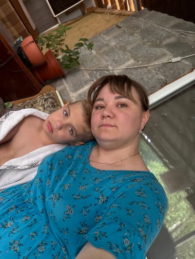 In a casual, indoor setting that appears to be a porch or covered outdoor area, a young boy and a woman are captured in a close-up, slightly tilted photograph. The boy, with blond hair and green eyes, is looking directly at the camera with a neutral expression. He is wearing a white towel draped around his shoulders. The woman, positioned behind and slightly above the boy, has brown hair and brown eyes. She is looking towards the camera with a gentle smile. She is wearing a blue dress with a floral pattern.
The background features a textured grey wall, possibly made of cinder blocks, with some wires and an electrical outlet visible. To the left, there is a wooden structure and a plant in a terracotta pot. A window or glass door is visible in the upper part of the frame, reflecting greenery and the outdoors, suggesting it might be daytime. The overall atmosphere of the image is intimate and relaxed, conveying a sense of connection between the two individuals. No visible text is present in the image. The location context of Nizhne-Chufichevo, Russia, provides geographical information but is not directly discernible from the visual elements of the photograph.