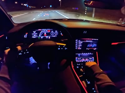 The image captures the interior of a car at night, as evidenced by the illuminated dashboard, road lights, and dark surroundings. The driver's perspective shows the steering wheel, digital instrument cluster displaying speed and other metrics, and a central infotainment system. Red ambient lighting outlines parts of the dashboard and console, creating a modern ambiance. The road ahead is visible through the windshield, with lane markings and distant streetlights indicating a highway or well-lit road. The location is identified as Bezden, Bulgaria. No people are explicitly visible, but the image implies the presence of a driver and possibly a passenger. The focus is on the car's interior technology and the nighttime driving environment.