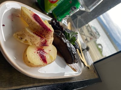 A plate of food features sliced potatoes topped with a vibrant red powder, and a portion of dark, glazed meat garnished with a sprig of parsley. A gold-colored fork and knife rest on a napkin to the side. In the background, a green can and a glass can be seen, suggesting a drink accompaniment. The scene is set near a window, with a glimpse of the outside visible.