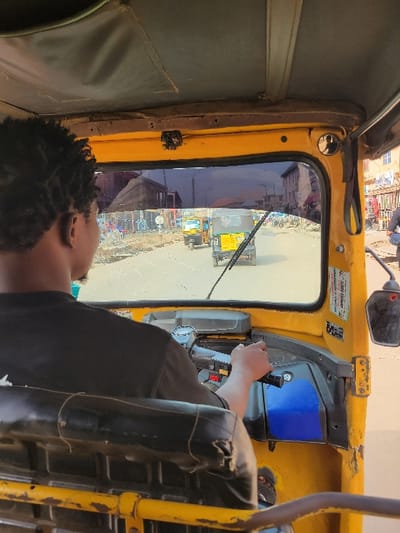 The image depicts the interior perspective from the driver's seat of a yellow motorized rickshaw traveling on a paved road in Puerto López, Colombia. A person with dark, curly hair, wearing a black short-sleeved shirt, is seated in the driver's seat, viewed from behind. Their right hand is on the black handlebar/steering mechanism. The rickshaw's interior features a worn black vinyl driver's seat with visible string or twine across its back, and a blue panel on the right side of the dashboard. The windshield appears tinted, and a single wiper blade is positioned in the lower center. An exterior rearview mirror is visible on the right.
Ahead, on the sunlit road, two other yellow motorized rickshaws are visible, traveling in the same direction. The road surface shows some irregularities, with faint white dotted lines. The surrounding environment consists of low-rise buildings, some appearing multi-story, lining both sides of the street. These structures exhibit varied architectural styles and signs of wear, housing what appear to be storefronts or market stalls. Several indistinct figures are present on the street and sidewalks. Overhead cables are visible in the distance. The overall scene indicates an active urban or semi-urban street during daylight hours.