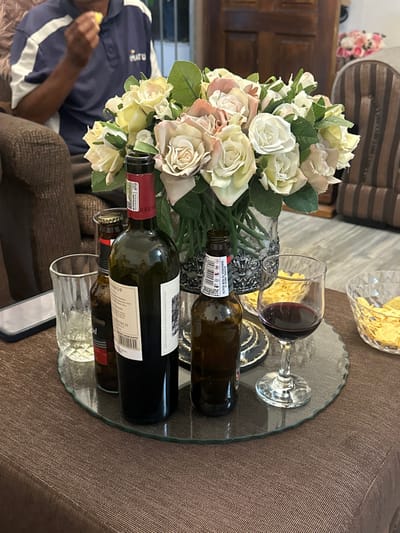 This image captures an indoor scene within a residence in Mitchells Plain, South Africa. The primary focal point is a round, scalloped-edge glass tray positioned on a dark brown, textured tablecloth. On the tray are: one uncorked bottle of Delheim red wine, one full, capped bottle of Hansa beer, one empty, uncapped bottle of Castle Lite beer, a stemmed glass containing approximately half a glass of red wine, and an empty clear tumblers-style glass. A large glass vase holding an arrangement of artificial roses in cream, pale yellow, and muted pink hues, accompanied by green foliage, occupies the center of the tray.

To the left of the glass tray, a smartphone with a dark case is placed on the tablecloth. To the right, two clear glass bowls contain potato chips. In the background, the upper torso and arm of an individual are visible, seated on a brown upholstered armchair. The person wears a navy blue polo shirt with white accents and is holding a small, light-colored object to their mouth. Further into the background, a wooden door or cabinet and a striped brown upholstered armchair are discernible.