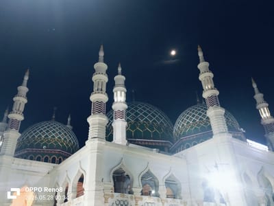 The image shows the Baitul Muttaqin Mosque in Kotapantonlabu, Indonesia, taken at night. The mosque features multiple green and gold domes with white latticework. The white exterior of the mosque is illuminated by strong artificial lights, creating a bright contrast against the dark night sky. Several tall, slender minarets with white and maroon accents rise from the mosque's structure. In the dark blue sky, a small, round light suggests the presence of the moon. At the bottom left corner of the image, there is a watermark indicating "OPPO Reno8" and a timestamp "2026.05.28 00:34". The perspective is from a low angle, looking upwards towards the mosque and the sky.