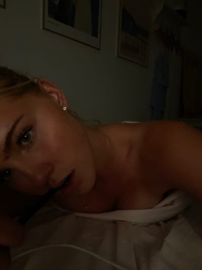The image depicts a dimly lit interior, characteristic of a private residence in Skanderborg, Denmark. No specific external geographical markers for Skanderborg are discernible within the frame. In the foreground, a young adult female, with light skin and blonde hair, is positioned on her side, resting on white bedding, likely a bed. Her face is angled towards the viewer, with her right eye visible and open, looking towards the camera. She is adorned with a small, clear stud earring in her right ear and a thin, gold-colored chain necklace with a small pendant. Her bare shoulders and upper chest are visible, partially covered by the white bedding. The background consists of a light-colored wall displaying two framed art prints; one appears to be a dark-toned landscape or abstract, and the other lighter. To the far right, indistinct shapes suggest hanging items, possibly articles of clothing. The overall lighting is low, indicating an evening or low-light indoor environment.