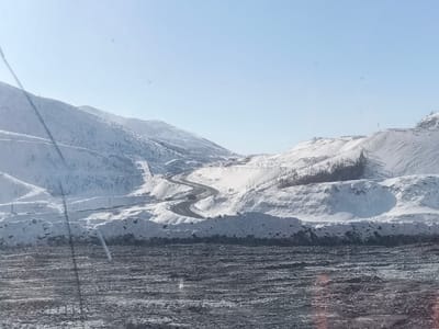 The image presents a wide-angle, panoramic view of a snow-covered, hilly, or mountainous landscape under a clear, light-blue sky, situated in the vicinity of the city of Im. Gastello, Russia. In the mid-ground, a dark, winding paved road traverses the snow-covered terrain, snaking through the hills. Scattered patches of dark, leafless trees are visible on the slopes, primarily to the right of the road. The background consists of multiple layers of snow-dusted peaks or large hills. The immediate foreground features a dark, uneven ground surface, appearing as exposed dirt, rock, or coal fragments, exhibiting irregular textures indicative of heavy vehicle traffic or excavation. No individuals, vehicles on the road, or active equipment are discernible. The scene is illuminated by daylight, characteristic of a clear winter day.