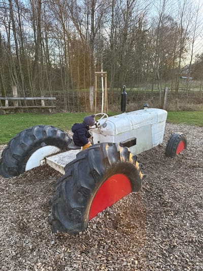 In Olingen, Luxembourg, an outdoor scene depicts a young child interacting with a static tractor playground structure. The child, dressed in a dark blue puffer jacket, yellow pants, and a grey knitted beanie, sits on a grey seat positioned on the tractor's frame. The child's hands are near a white steering wheel and a black lever. The tractor structure is an older model, painted light grey, with a black exhaust pipe visible on the right side of its engine cowling. It features oversized, real tractor tires embedded in the ground; the visible rear left tire has a white rim, the visible rear right tire has a red rim, and a smaller front tire also shows a red rim. The ground immediately surrounding the tractor is covered in dark brown wood chips. Beyond this area, green grass is visible, followed by a rustic wooden bench made of logs. A wire fence runs horizontally across the mid-ground, separating the immediate play area from a dense background of numerous bare deciduous trees under a bright, possibly overcast, sky. A young, staked sapling stands near the tractor, and faint sunlight penetrates the trees in the distance.