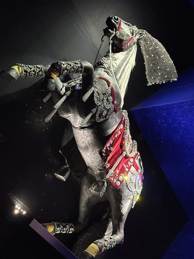 The image shows a highly ornate, life-sized sculpture of a horse, dramatically lit against a dark background with a blue, angular structure to the right. The horse is in a rearing pose, with its front legs raised. It is covered in a multitude of embellishments, including sparkling rhinestones and intricate silver detailing. The horse wears a red and gold draped saddle or cloth across its body, also heavily adorned with jewels. A mesh-like, crystal-encrusted veil or mane extension hangs from its head. The sculpture is on display in an exhibition space, likely within the Swarovski Kristallwelten (Crystal Worlds) in Wattens, Austria, given the context of elaborate crystal displays and the architectural style visible. The lighting is dramatic and theatrical, focusing attention on the detailed artistry of the horse sculpture.