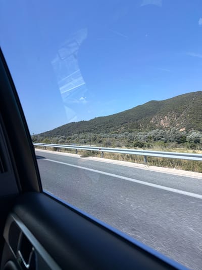 The image captures a view from inside a vehicle, looking out a window onto a highway. The road is paved with grey asphalt and has a white line marking the lane. To the right of the road, there is a metal guardrail separating the highway from a grassy embankment. Beyond the embankment, a body of water is visible, bordered by dense vegetation and olive trees. Further in the background, a large, verdant hill slopes upwards, covered in trees and some rocky outcrops. The sky is a clear, bright blue with a few faint contrails visible. Reflections of the car's interior, including a rearview mirror, are visible on the window. The location is identified as a part of the Municipal Unit of Promachonas in Greece.