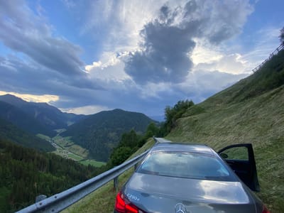 A gray Mercedes-Benz C200d sedan is parked on the side of a winding road in a mountainous valley, with the driver's side door open. The car is facing away from the camera, with its rear end visible. The road curves around a grassy hillside, which is bordered by a metal guardrail. In the background, a lush green valley stretches out, dotted with houses and surrounded by dense pine forests. The valley is framed by imposing, dark mountains under a dramatic sky filled with heavy, dark clouds and patches of bright sunlight.

The time of day appears to be late afternoon or early evening, as indicated by the low angle of the sun and the interplay of light and shadow. The weather is dynamic, with stormy clouds suggesting a recent or impending rain shower. The location is Ratschings - Racines, Italy, a region known for its alpine scenery. The overall mood is one of adventure and natural beauty, with a hint of the dramatic power of nature. The visible text on the car reads "C200d" and the Mercedes-Benz logo.