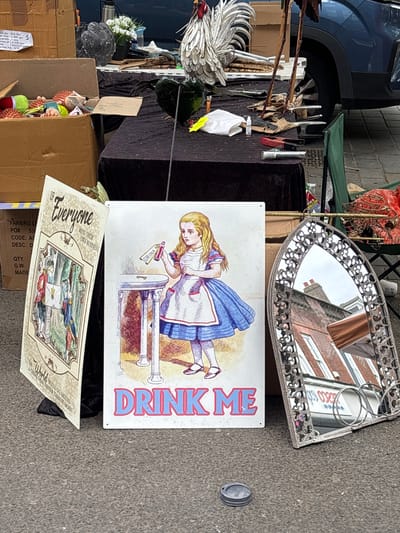 The image shows a collection of items arranged on the ground in what appears to be an outdoor market setting. Dominating the view is a white sign with a vintage-style illustration of Alice from "Alice in Wonderland," labeled "DRINK ME." Beside it is another sign with the text "Everyone Mind Their Own Business." An ornate, silver-framed mirror reflecting surrounding buildings lies nearby. These items are amidst boxes and other miscellaneous goods, suggesting a flea market or similar sales environment. A blue car can be seen on the left of the frame and a building reflected in the mirror, both suggesting this is an outdoor market or boot sale in St Albans, United Kingdom.