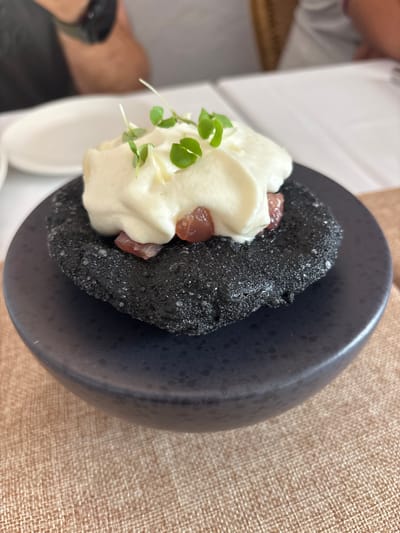 A close-up shot focuses on a small, dark, circular pastry topped with a dollop of white cream and garnished with microgreens. The pastry appears to be made from a dark flour or dough, possibly charcoal-infused, with a slightly rough texture. Beneath the cream, small pieces of red, finely diced food, likely tuna or another type of fish, are visible. The entire dish is presented on a dark, shallow plate, which sits on a textured, light brown placemat. In the blurred background, two individuals are seated at a table with white tablecloths, suggesting a dining setting. One person on the left is wearing a black t-shirt and appears to be looking down. The other person on the right is wearing a light-colored shirt. The setting is presumed to be in Carboneras, Spain, a coastal town known for its seafood restaurants, which would be consistent with the presentation of a dish featuring raw fish.