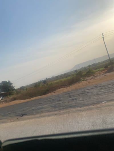 A slightly hazy daytime landscape features a road's edge in the foreground, transitioning into fields and distant hills. Utility poles with wires extend into the sky. The left side of the image is predominantly a clear blue sky, contrasting with the earthier tones of the landscape. A dark object is in the corner of the frame.