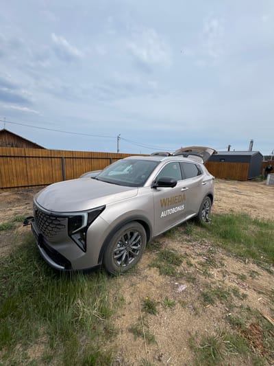 A light-colored SUV is parked on a dirt road with patchy grass. The car's trunk is open. Behind the car, a wooden fence runs horizontally across the image. Further back, there are several small structures, including a dark-colored building with a metal roof and a chimney. The sky is overcast. A power pole with wires is visible to the right of the wooden fence. The scene appears to be in a rural or semi-rural area, consistent with Khuzhir, Russia, known for its natural landscapes and remote setting on Olkhon Island.