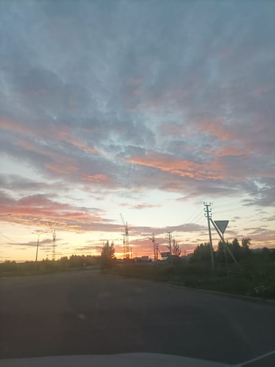 The image captures a sunset scene in Zapadny, Russia, with a road leading into the distance. The sky is filled with dramatic clouds painted in hues of pink, orange, and grey, indicating the sun is setting. Several construction cranes are visible on the horizon, suggesting ongoing development or building projects in the area. Electrical poles with wires crisscross the sky, and a triangular road sign is prominently placed on the right side of the image. Trees and other foliage line the sides of the road, with some buildings faintly visible in the distance. The overall mood is tranquil and slightly dramatic due to the striking sunset colors. The time of day is clearly evening, and the weather appears to be clear, allowing for a vibrant sunset. There is no visible text in the image.