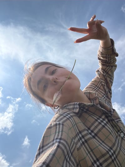 A young woman, with dark hair and brown eyes, is captured from a low-angle perspective, looking up at the sky. She is wearing a tan and brown plaid shirt. A long, thin stem with a fluffy, pinkish flower is held between her teeth. Her left hand is raised, with fingers spread in a gesture, against a bright blue sky with scattered white clouds. The scene appears to be outdoors, possibly in a grassy or field-like area, given the presence of the plant. No specific city or location details are visible in the image.