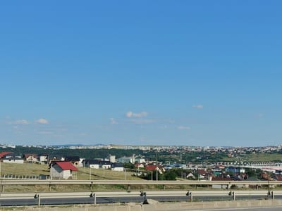 The image captures a panoramic view of a suburban or semi-rural area near Șcheia, Romania, under a clear blue sky with sparse clouds. In the foreground, a highway guardrail separates the viewer from a road surface. Beyond the guardrail, a gently sloping landscape is dotted with numerous residential buildings, predominantly houses with various roof colors, including red, brown, and black. Some houses appear to be under construction, with unfinished exteriors. A fence runs along a portion of the property line.

In the middle ground, the cluster of houses gives way to a denser area of buildings, with some appearing to be larger structures, possibly commercial or institutional, interspersed with more houses. The terrain continues to undulate, with trees and vegetation covering the hillsides.

The background reveals a more expansive cityscape stretching across the horizon. This distant urban area features a concentration of multi-story buildings, indicative of a town or city center. The overall impression is one of a developing community, with a mix of residential construction and established urban areas. The natural environment is characterized by rolling hills and abundant greenery. No people are visible in the image.