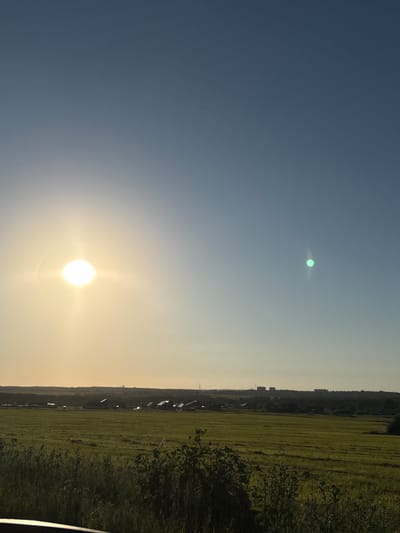 The image depicts a vast, clear blue sky transitioning to a lighter yellow hue near the horizon. The sun, a bright white orb radiating light, is positioned in the upper left quadrant. To the right of the sun, a smaller, luminous green circular object is visible. In the distance, a flat landscape stretches across the lower portion of the image, characterized by a large, green field. A line of trees and buildings forms a dark silhouette against the sky along the horizon, with a few taller structures, likely apartment buildings, standing out. The foreground is dominated by tall grass and some foliage. The scene appears to be a rural or semi-rural area.
