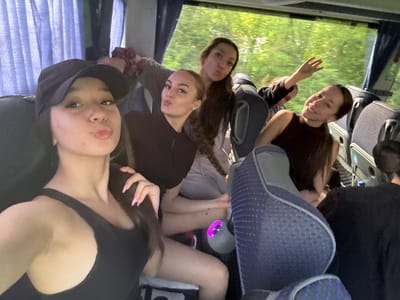 The image displays the interior of a passenger bus or coach occupied by five individuals, predominantly young women, during daylight hours. Dense green foliage is visible through the large windows, suggesting transit through a natural environment. In the immediate foreground, on the left, a young woman wearing a black baseball cap and a black tank top is captured in a selfie-style shot, pursing her lips. Her right hand, featuring pink iridescent manicured nails, is visible in the lower left quadrant. To her right and slightly behind, another young woman with dark hair styled in a braid also makes a pursed-lip expression, wearing a dark long-sleeved garment. Behind them, a third young woman with dark hair and a light-colored top adopts a similar facial expression. Further right, a fourth young woman with dark hair, wearing a dark sleeveless top, smiles and has her left arm raised with fingers splayed. The back of a fifth person's head and dark-clad shoulder are partially visible on the far right. The bus interior features blue and grey patterned seating, blue window curtains pulled back on the left, and a single illuminated purple light ring near the floor between seats. There is no discernible visual information within the image to specify the location as the city of Boichovtsi, Bulgaria.