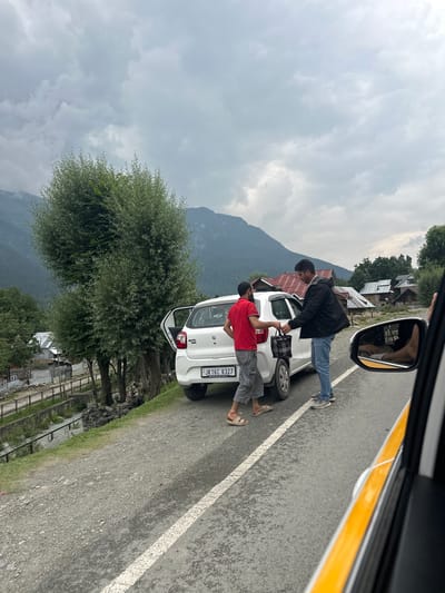 In the city of Ravil, India, a man in a red t-shirt and grey pants hands a patterned bag to another man in a black jacket and blue jeans. They stand beside a white car with its door open on the side of a road. The car has a license plate that reads "JK16C 8327". In the background, there are trees, houses with red roofs, and mountains under a cloudy sky. A side-view mirror of another vehicle is visible in the foreground on the right.