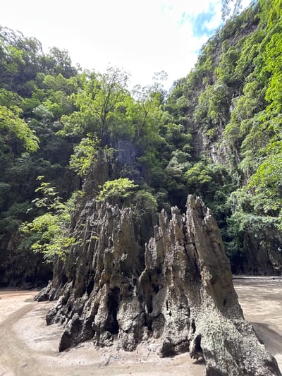 This image shows a limestone karst formation on a sandy beach in Krasom, Thailand. The formation is tall and jagged, with sharp spires and rough, textured surfaces. Lush green vegetation covers the surrounding cliffs, which rise steeply on either side of the beach. The sky above is bright, with some clouds visible. The scene appears to be a natural landscape, possibly a tourist attraction given its photogenic qualities. There are no people or any signs of human activity other than what appears to be a path on the sandy ground.