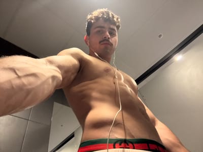 A low-angle indoor photograph depicts a muscular young male with light skin, dark curly hair, and a mustache, positioned in Chantepie, France. He is shirtless, wearing white boxer briefs featuring a green and red waistband with a "GG" logo. White wired earbuds are inserted into his ears, with the cable extending down his chest. His right arm is extended towards the camera, showcasing prominent muscle definition and visible veins, suggesting he is taking a selfie. The subject looks directly forward with a neutral expression. The background features dark grey tiled walls on the left and lighter, possibly reflective, walls on the right. A white ceiling with a circular recessed light fixture is visible, indicating artificial illumination of the scene.