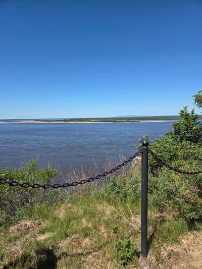 The image captures a panoramic view overlooking a vast body of water, likely a wide river, under a clear blue sky. The perspective is from an elevated position, looking across the water towards distant land.

In the foreground, on the right side, there is a grassy embankment covered with a mix of green and drier vegetation, including bushes and small trees. A prominent feature is a sturdy black metal post with a spherical finial, located in the mid-right. A heavy black metal chain extends from this post, curving downwards and out of the frame towards the bottom right, suggesting it forms part of a protective barrier or fence along the edge of the embankment.

The wide river stretches horizontally across the middle of the frame, its surface appearing calm with a deep blue or grey hue. On the far side of the river, a thin strip of lighter-colored land, possibly a sandbar or distant shore, is visible, backed by a greener, forested area. Faint outlines of low mountains can be seen on the horizon in the far distance, blending into the sky.

The left half of the image is dominated by an expansive, cloudless bright blue sky, indicative of a clear and sunny day. The overall scene conveys a sense of tranquility and vastness. There is no specific activity or event taking place, and no people are visible. There is no visible text in the image. The setting appears to be a natural landscape, possibly a viewpoint overlooking a major waterway, consistent with the location context of Jiejinkou, China.