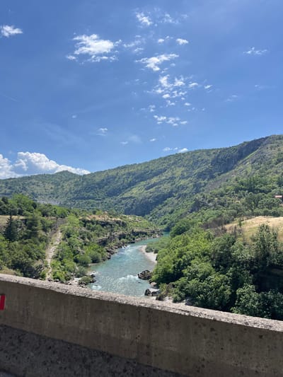 A daytime, landscape view from Donje Mrke, Montenegro, features a river running through a valley at the base of a large, tree-covered mountain. The blue river has areas of rapids and some visible rocks. Lush, green trees and vegetation cover the lower portion of the mountain and line the river's banks. A tall, green mountain rises to the left of the river valley. The top of the mountain features a patch of tan-colored dry grass and a small building. A concrete structure is visible in the foreground. The sky is bright blue with scattered white clouds.