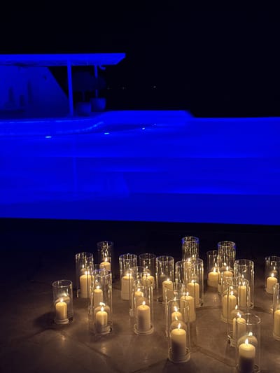 The image depicts a scene in Ghadeer Al Tayr, United Arab Emirates. On the left side, a structure illuminated with bright blue lighting is visible. This structure has a multi-tiered design. To the right, a dark wall is adorned with numerous glass cylinder candle holders, each containing a lit candle. The background is dark. The scene gives the impression of a contemporary architectural element combined with decorative lighting.