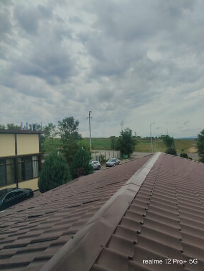 The image displays a view from a brown tiled roof overlooking a landscape with a building, trees, cars, and a cloudy sky. In the foreground, the corrugated texture of the roof tiles is prominent. To the left, a beige building with dark windows is visible, with a black car parked in front of it. Behind the building, several green coniferous trees stand, and further back, deciduous trees are scattered across a green, hilly terrain. Two silver cars are parked on a paved area. Electrical poles and wires are present in the mid-ground. The sky is overcast with a mix of light and dark gray clouds. The text "realme 12 Pro+ 5G" is visible in the lower right corner of the image. The scene appears to be set in an outdoor, semi-rural or suburban environment, possibly on the outskirts of a city.