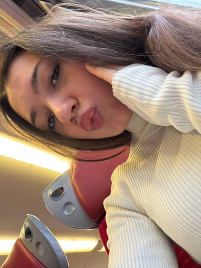 A young female is depicted in a selfie, taken within what appears to be a passenger compartment of a train or bus, situated in Obermodern-Zutzendorf, France. The subject, positioned centrally and tilted, has long, dark brown hair falling across her shoulders and upper back. She is wearing a light-colored, ribbed turtleneck sweater. Her left hand is raised to her left cheek, gently pressing against it, while her lips are deliberately pursed in a "duck face" expression. Her eyes are open and directed towards the camera. The background reveals portions of red upholstered seating with distinctive grey plastic armrest components. Overhead, elongated light fixtures emit a warm, artificial illumination across the interior. A distinct reflection of a smartphone's camera lens is visible on the grey plastic surface of a seat armrest in the mid-left, indicating the device used to capture the image.