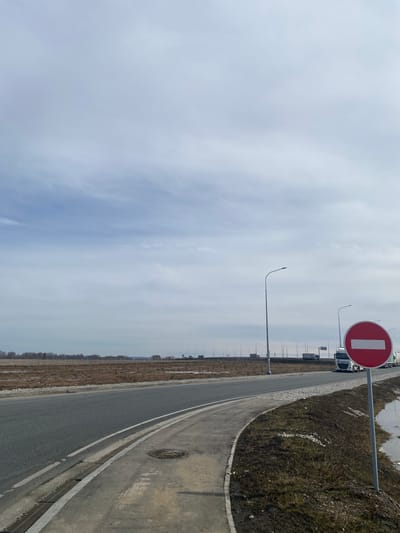 A road in the vicinity of Zarya, Russia, is depicted on an overcast day. The road is paved, with a white line marking the edge. A red and white "no entry" sign is prominent on the right side of the road. Streetlights line the road. Beyond the road, there is a patch of land with sparse vegetation. A series of vehicles, mainly trucks, are parked in the distance.