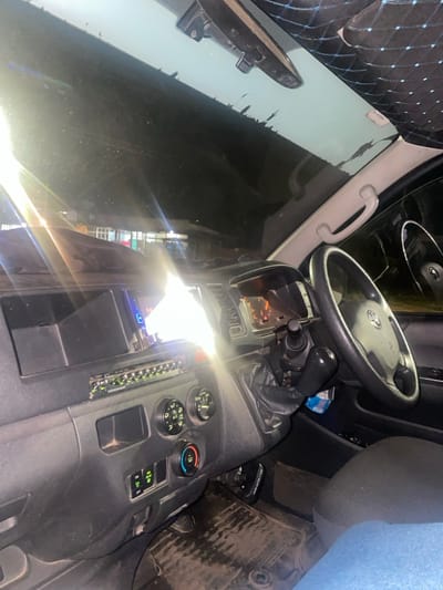 The image displays the interior of a vehicle, looking forward through the windshield at night in Kiaga, Kenya. The primary focus is the dark grey dashboard and controls. A black, three-spoke steering wheel with a Toyota emblem is positioned on the right side, indicating a right-hand drive configuration. Behind the steering wheel, the instrument cluster is illuminated with amber-orange light, suggesting the vehicle is operational. A black manual gear shift lever extends from the dashboard's center.
To the left of the steering column, an aftermarket infotainment screen emits a bright white light, below which an audio equalizer panel with multiple green illuminated indicators is visible. Below these are two circular climate control dials and a row of four green-illuminated rectangular switches. The floor beneath the dashboard is covered with dark, textured mats, showing signs of wear. A segment of a human leg, clad in blue denim, is visible in the lower right corner, indicating a passenger.
Through the windshield, a dark nighttime street scene is visible. Distant buildings with illuminated windows or facades are discernible against the darkness. Multiple bright light sources outside the vehicle create significant lens flare across the dashboard and windshield. The rearview mirror is affixed to the top center of the windshield, and a quilted black fabric headliner with blue stitching is partially visible at the top right. The upper edge of the windshield exhibits a black, irregular strip that appears to be peeling or damaged.