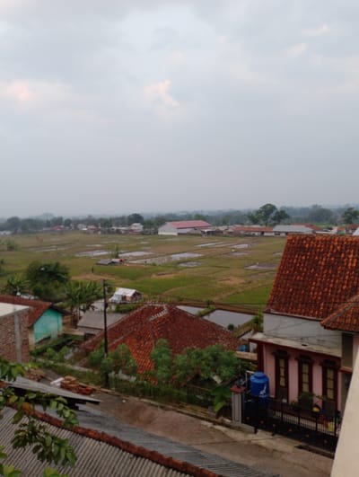 This is a high-angle, full shot of a rural landscape on an overcast day in Sukamanah, Indonesia. In the foreground, there are several houses with terracotta tile roofs. One house on the right is painted pink with dark brown window frames and a black metal fence. A blue water tank is visible next to this house. To the left, a house with a light green exterior wall is partially visible. Beyond the houses, there are extensive rice paddies, some of which are flooded. Further in the distance, more houses and trees are scattered across the landscape, with a hazy mountain range visible on the horizon. The sky is uniformly cloudy and bright white.
