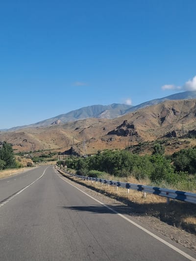 A road curves into the distance, flanked by a metal guardrail and dry grass. Beyond the road, the landscape rises into arid, rocky hills and mountains under a clear blue sky. Power lines are visible on the hillsides, and some sparse vegetation dots the terrain. The scene appears to be in a rural or mountainous area of Armenia.
