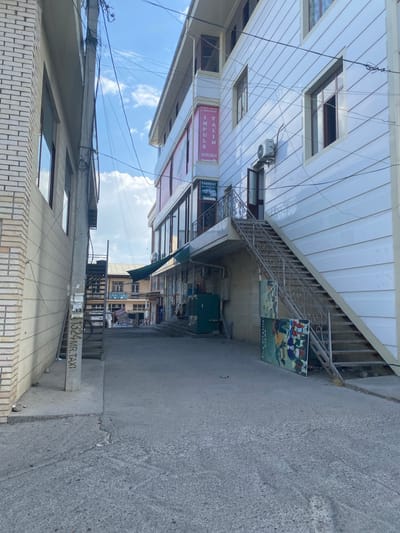 This is a full, eye-level, outdoor shot of a narrow street in Parkent, Uzbekistan, featuring a multi-story building with an exterior staircase on the right and a building with a brick facade on the left. The sky above is blue with scattered white clouds. The street is paved with asphalt and shows some cracks and debris.
On the right, a white building with horizontal siding has several windows. A metal staircase with an ornate railing ascends along the side of the building. An air conditioning unit is mounted on the wall. Below the stairs, there is a green metal enclosure and a large, colorful advertisement board. A sign for "PARKENT AVTOMAKTABI" (Parkent Driving School) is visible on the building.
On the left, a building with a light-colored brick facade has large windows. A utility pole with numerous wires runs vertically between the two buildings. A set of outdoor stairs made of concrete and metal leads up to a higher level. A pole with graffiti and a bicycle wheel leans against it. Further down the street, more buildings with shops are visible, with awnings and signs.
There are no people visible in the frame. The scene appears to be a typical urban streetscape, possibly a commercial area, with a slightly neglected appearance due to the debris on the ground and the condition of the utility pole.