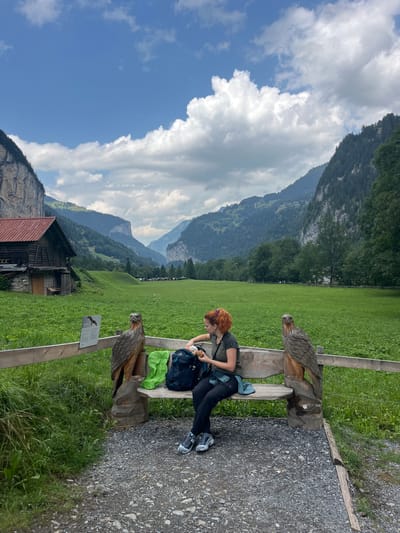 A woman with red hair is sitting on a wooden bench in Lauterbrunnen, Switzerland. She is looking down at a backpack at her feet and holding a piece of food. The bench is carved to look like an eagle on either side. Behind her, a large green field stretches towards mountains. To the left of the field, there is a wooden building with a red roof. The sky is blue with some clouds.