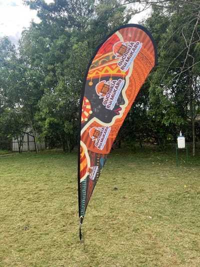 This outdoor image, captured in Makuyu, Kenya, features a vibrant teardrop-shaped banner prominently displayed on a green lawn. The banner, oriented vertically due to the image's sideways capture, is adorned with rich orange, brown, and black hues, featuring intricate geometric and traditional patterns.
The primary text repeated across the banner is "GuChokia Rui MUKARO" and "a Kikuyu Culture Podcast," indicating that the banner is promoting a podcast focused on Kikuyu culture. A stylized logo, possibly depicting a traditional vessel or bowl, is also visible alongside the text.
In the background, lush green trees provide a natural canopy, and a patch of sky, appearing overcast or partly cloudy, suggests it is daytime. Further back, nestled among the foliage, a small white box on a green metal stand is visible, possibly a hand sanitizer station or an environmental sensor. The setting appears to be a verdant, open area, possibly for an event or public display. No individuals are present in the frame.