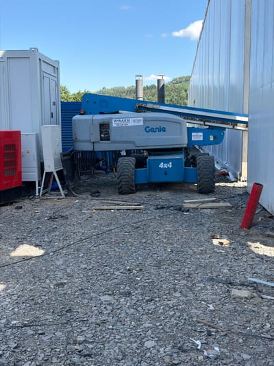 The image depicts a construction site in Janowice, Poland. In the foreground, a gray and blue Genie boom lift is positioned on a gravel surface. The boom lift has "4x4" marked on its base. To the left of the boom lift, a red generator is visible, along with a white electrical panel. In the background, a large white building with a corrugated metal facade dominates the scene. The sky is blue with a few white clouds. The ground is covered in gravel, with some scattered debris and planks. There are no people visible in the image.