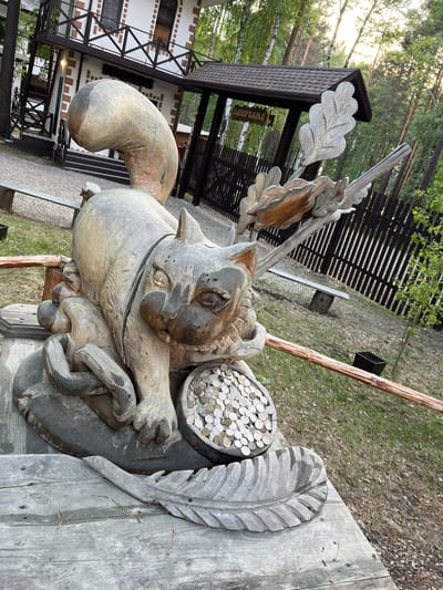 The image shows a carved wooden sculpture of a cat sitting in front of a building in Tal'tsy, Russia. The cat is large and detailed, with coins piled in a bowl-like structure in front of it. The building in the background appears to be a traditional Russian wooden house with a pitched roof and a balcony. The surrounding environment is a wooded area with a fence and benches. The sculpture is placed on a wooden platform, with a carved feather in the foreground. The overall scene suggests a tourist attraction or a cultural site within the village.