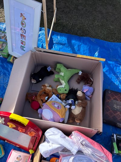 The image shows a collection of items arranged on a blue tarpaulin. Dominating the view is a cardboard box filled with stuffed animals, including teddy bears, a green dinosaur, and a black dog. Adjacent to the box is a framed picture with the name "Joseph" displayed in colorful, animal-themed letters. To the right of the box, there is a bag labeled "M&S SCHOOL," and several toys, including a red and yellow magnetic drawing board. The objects suggest a yard sale or flea market setting. Based on the OCR text in the image, it's highly likely that the image was taken in Chirk, United Kingdom.