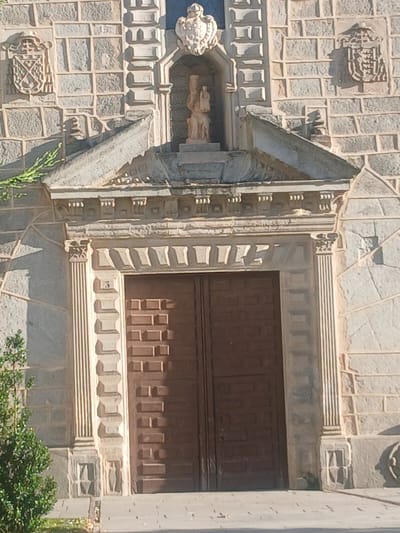 The image shows the ornate entrance to a building, likely a church or historic structure, with a large wooden double door as the central focus. The door is made of dark wood and features a raised, patterned design consisting of rectangular blocks. Flanking the door are fluted pilasters, which are decorative columns with vertical grooves. Above the door, there is a stone lintel with intricate carvings and decorative elements. Further up, a triangular pediment juts out, framing a niche that contains a statue of the Virgin Mary and child. The facade of the building is constructed from stone blocks, with decorative shields mounted on either side of the upper section. A small amount of greenery is visible to the left of the entrance. The lighting suggests it is daytime, with shadows cast by the architectural features. This building is identified as the Church of San Vicente in Ávila, Spain.