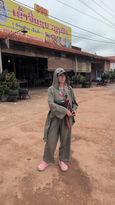 The image depicts a young woman standing on a dirt road in front of a building with a large yellow sign. She is wearing a hooded gray coat, a t-shirt, and pink crocs. She has blonde curly hair and appears to be holding a cigarette. The building behind her has a tiled roof and a sign with Thai or Laotian script, indicating a shop, possibly a bakery given the images of bread on the sign. The sign also contains numbers and phone numbers. The background shows more buildings and some vegetation. The weather appears overcast. The overall scene suggests a casual outdoor setting in a town or village.