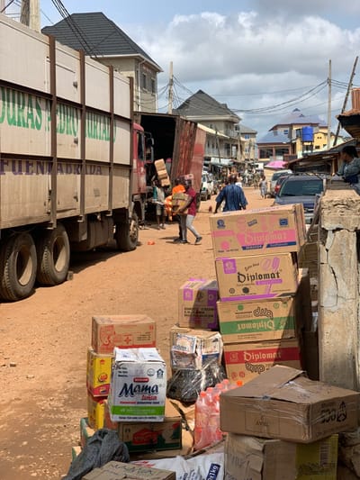 The image shows a street scene in Ondo, Nigeria, located at the coordinates 7.251 latitude, 5.196 longitude. A large white truck is parked on the left side of the street, partially blocking the view of the road. The truck has the words "DURAS PEDRO MURTAD" and "FUENTE ABRADA" written on the side in large green letters. There are several cardboard boxes stacked next to the truck. Several men are in the scene, including one man dressed in a bright red shirt standing in front of the truck and another man in a blue shirt standing in front of the boxes. There are several other men standing or walking in the background. The scene is characterized by a cluttered, busy environment with a mix of buildings and vehicles. The ground is dirt. The sky is blue and partly cloudy.