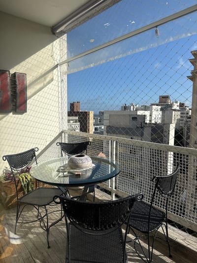 This image presents a sunlit residential balcony overlooking a dense urban cityscape in Porto Alegre, Brazil. The balcony is enclosed by a fine-mesh safety net and features a clear roll-up screen at its upper perimeter. Furnishings include a round glass-top table with a dark metal base, surrounded by four matching dark-colored chairs with woven seats and backs. Centered on the table, a light-colored ceramic bowl adorned with dark, abstract patterns rests on a white doily, accompanied by a small red box and a small circular object. Two rectangular art pieces, displaying abstract red and black designs, are mounted on the light-colored wall to the left. A wooden planter containing trailing green succulents is visible on the weathered wooden plank floor. Beyond the balcony, numerous multi-story buildings dominate the horizon under a clear blue sky. No individuals are present.