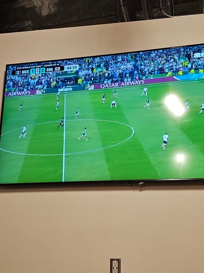 The image shows a large television screen displaying a live broadcast of a soccer match. The score on the screen indicates that Mexico is playing England, with the score at 1-2, and the time is 44:39. The game is part of the FIFA World Cup 2026, Round of 16. The field is green, and players in white and green uniforms are visible on the field. The stadium is full of spectators. There are advertisements visible, including "QATAR AIRWAYS" and "MEXICO CITY." The television is mounted on a wall, and a power outlet is visible on the wall below the screen. The scene appears to be in a room with neutral-colored walls. There is no visual information in the image that would indicate the location is the city of Seattle, United States of America.