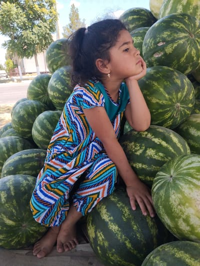 In Kiyat, Uzbekistan, a young girl with dark, curly hair tied in a ponytail sits amidst a large pile of watermelons. She is wearing a colorful, geometric-patterned dress with a teal neckline and short sleeves. Her right arm is bent, and her chin rests on her hand as she gazes thoughtfully to the right. Her bare feet are visible at the bottom of the frame, resting on a wooden surface. The watermelons are large and round, with dark green rinds and lighter green stripes. In the background, trees with green foliage and a building are visible under a bright, cloudy sky. The scene appears to be outdoors, possibly at a market or roadside stand.