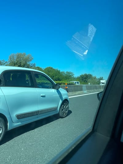 The image shows a light blue Renault Twingo driving on a highway. The location may be near Roccasecca, Italy, based on the environment along the highway, which includes trees and shrubs. Other vehicles, including a white van and a white truck, are visible in the distance on the same highway. The image appears to have been taken from inside another vehicle. The sky is clear and blue.